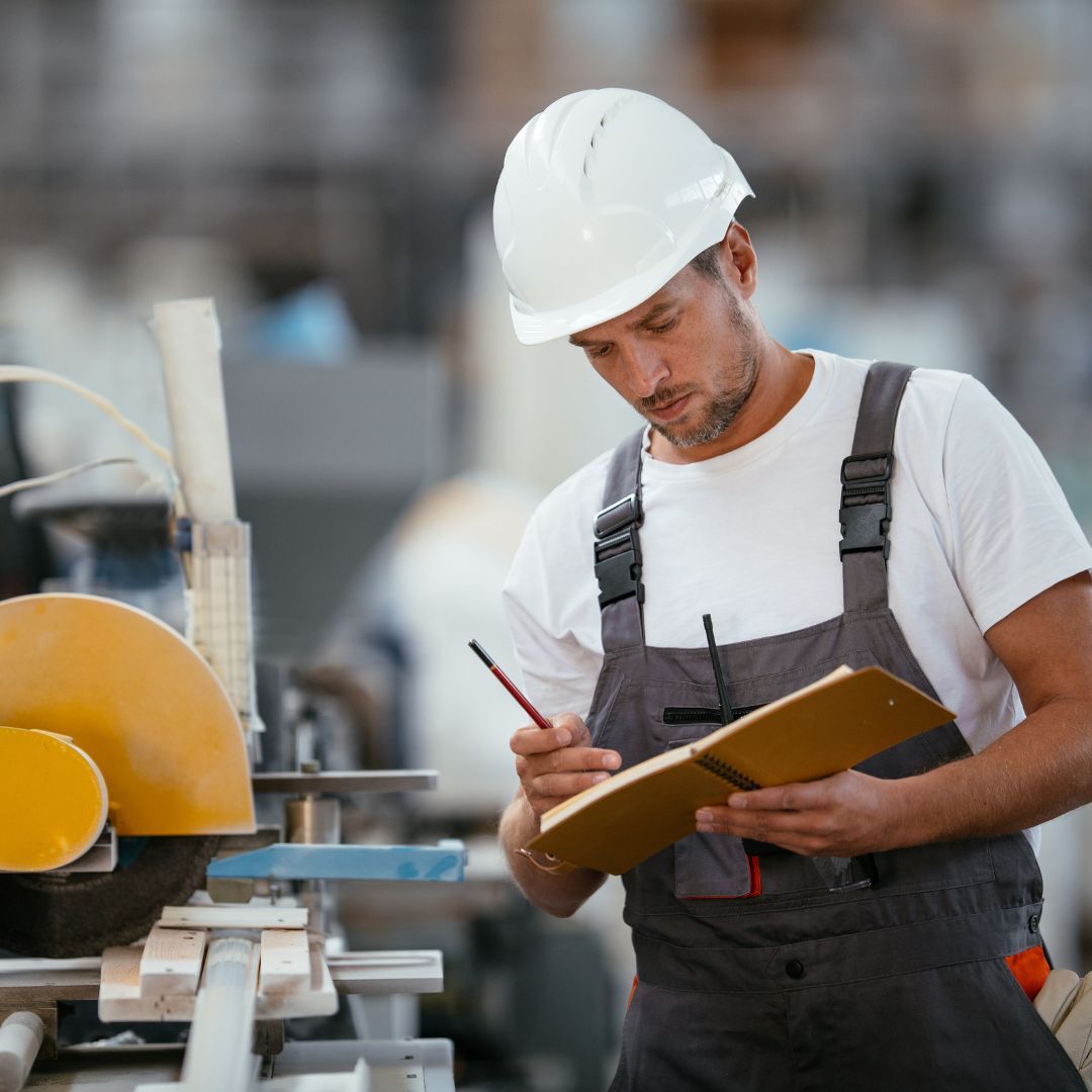 Technicus in overall en veiligheidshelm noteert bevindingen op een klembord tijdens een asset-inspectie in de werkplaats.